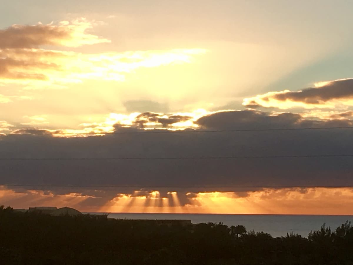 A stunning sunset is captured, with rays of sunlight breaking through the clouds and illuminating the horizon. The ocean is visible in the distance, reflecting the warm colors of the sky. Silhouettes of nearby vegetation add depth to the serene landscape.