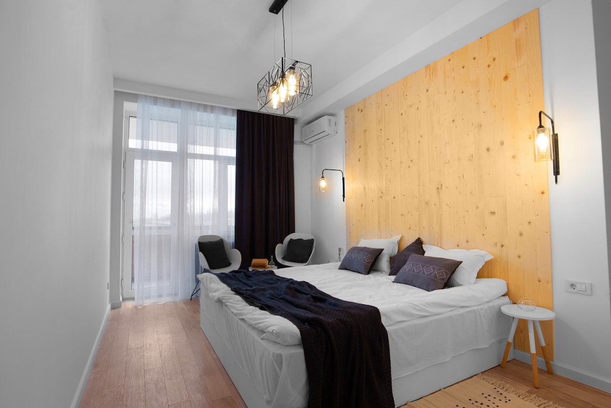 A bright sleeping area features a spacious double bed with white linens and decorative pillows. Two modern chairs are positioned beside a large window draped with sheer curtains, while a wooden accent wall adds warmth to the minimalist decor.