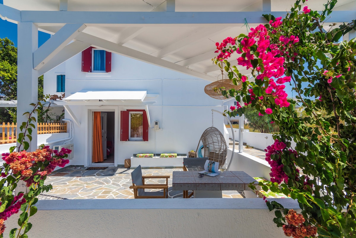 A charming outdoor space features a stone patio surrounded by vibrant bougainvillea. A suspended chair offers a relaxed seating option, while a wooden table and chairs are positioned for outdoor dining. The entrance to the house is framed by bright red shutters.
