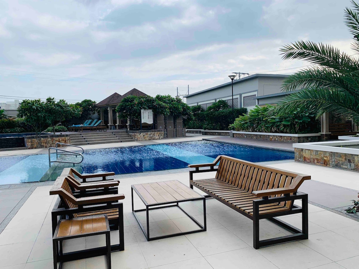 A modern outdoor area features a large swimming pool surrounded by lush greenery. Comfortable seating arrangements, including several wooden benches and a table, are positioned nearby, offering a relaxing space to enjoy the view. The setting provides ample sunlight and a tranquil atmosphere.