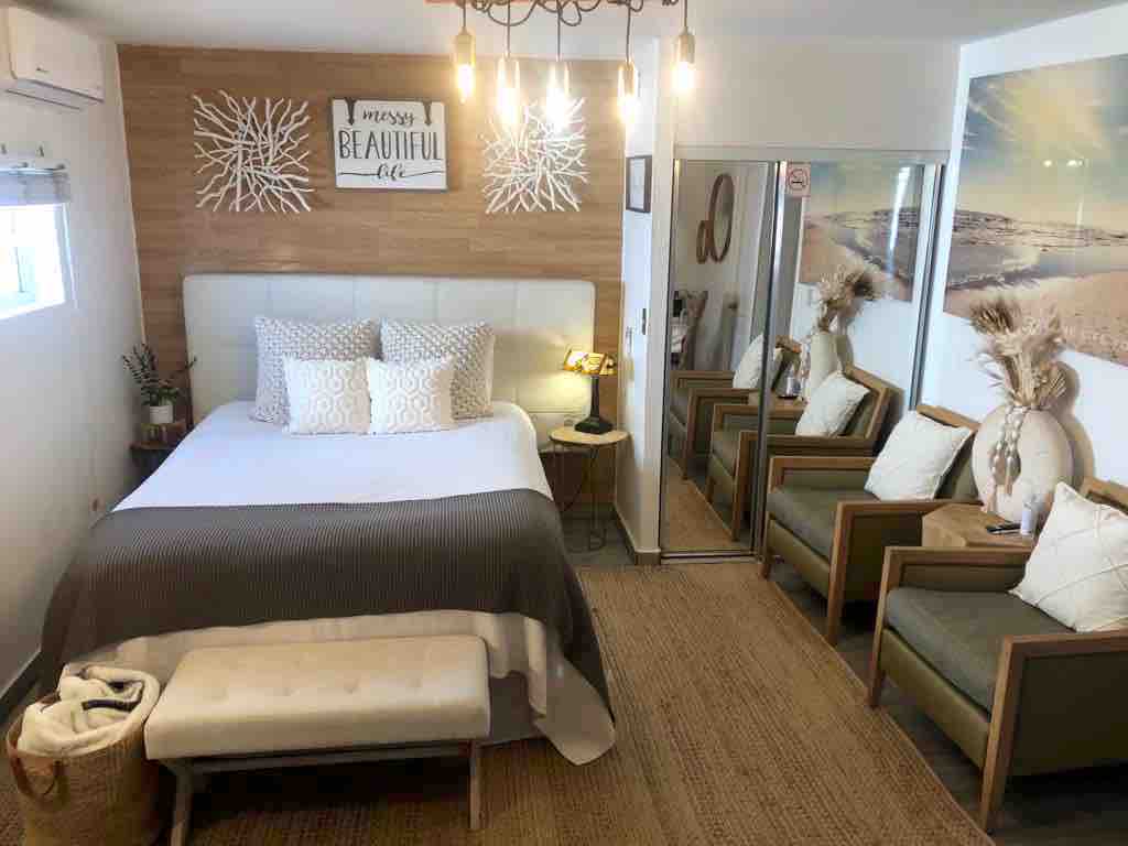 A studio space is shown, featuring a queen bed dressed in white linens and decorative pillows. A small bench sits at the foot of the bed, with two comfortable armchairs positioned beside it. A mirror reflects the light from nearby lamps, with wall decor adding a subtle touch.