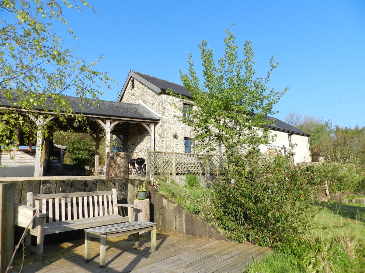 A stone cottage is pictured against a clear blue sky. The structure features a blend of traditional architecture and surrounding greenery. A wooden bench sits on a deck in the foreground, while trees and shrubs provide a natural setting, offering an inviting outdoor space.