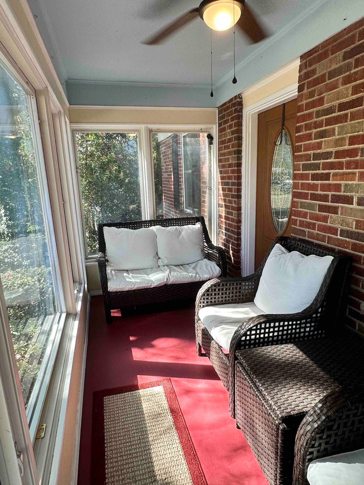 A sunlit enclosed porch features two wicker chairs with cushioned seats, framed by large windows that provide a view of the surrounding greenery. A decorative rug and overhead ceiling fan add to the cozy ambiance.