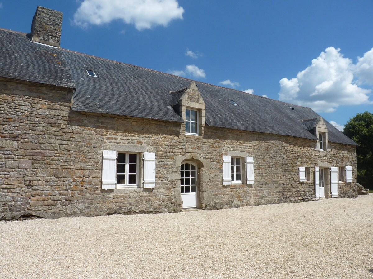 A historic stone building is displayed under a bright sky, featuring large windows with white shutters. The entrance is highlighted by a central doorway, providing a welcoming appearance. The surrounding gravel area contributes to the property's ample outdoor space.