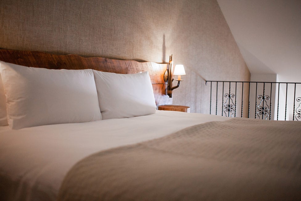 A cozy sleeping area is presented with a spacious bed topped with soft, white linens. The natural wooden headboard adds warmth against the textured wall. Ambient lighting is provided by a wall-mounted sconce, enhancing the room's tranquil atmosphere.