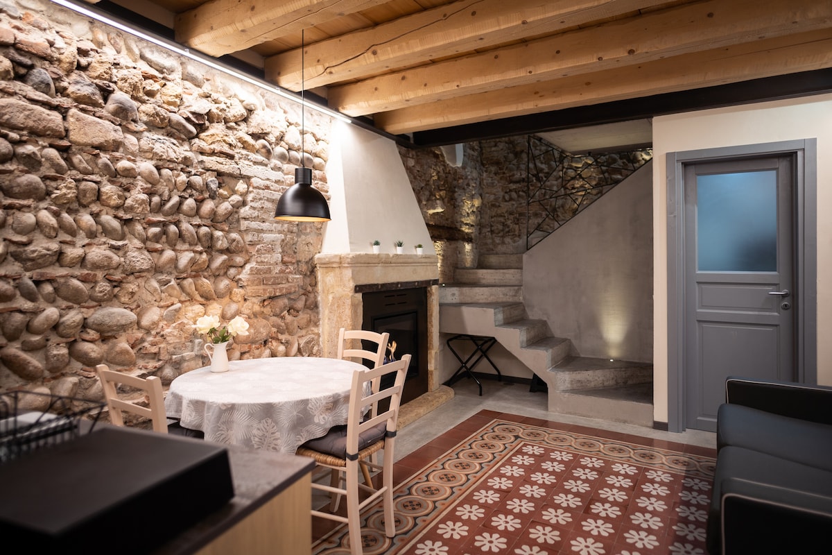 A rustic living space showcases an exposed stone wall, adding historical character. A round dining table with four chairs is positioned beside a gas fireplace. The floor features patterned tiles, while a staircase leads to the upper level, illuminated by soft lighting.
