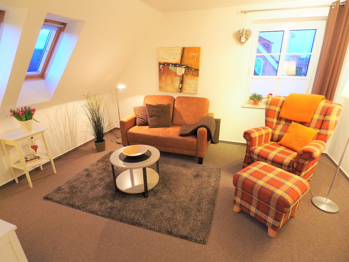 A cozy living area features a brown sofa with soft cushions and an orange plaid armchair. A round coffee table sits atop a plush area rug, with a floor lamp providing warm light. Natural light comes through a window, enhancing the inviting space.