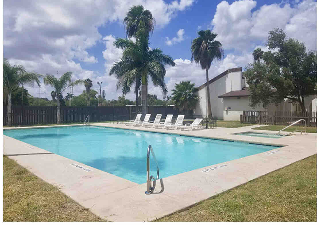 A clear blue swimming pool is framed by palm trees and green grass. Several white lounge chairs are positioned around the poolside, offering a relaxing space to unwind. The surrounding area is enclosed by a wooden fence and features well-maintained landscaping.