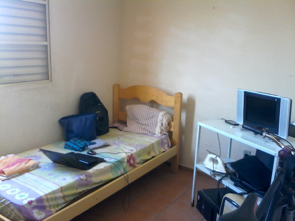 The image displays a simple bedroom featuring a wooden bed with a patterned blanket. A small desk holds a monitor and a laptop, while a chair and backpack are positioned nearby. Natural light enters through a window with horizontal blinds, illuminating the space.