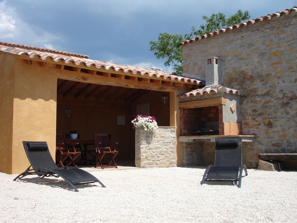 An outdoor area features a stone barbecue, shaded seating for dining, and two lounge chairs positioned on a gravel surface. A wooden pergola enhances the space, which is surrounded by rustic stone walls and greenery in the background.