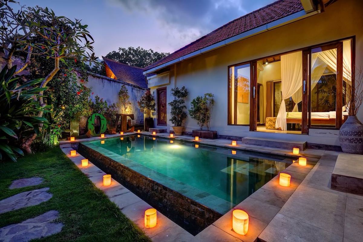 An inviting private pool glows softly, surrounded by decorative lanterns on the stone patio. Lush greenery frames the scene, with tropical plants and trees enhancing the tranquil environment. The villa's open living space is elegantly designed, inviting relaxation under the evening sky.