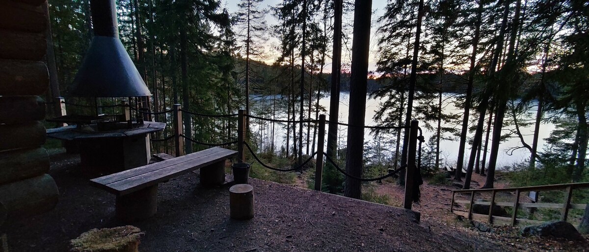 A serene outdoor space is framed by tall trees, with a view of a calm lake reflecting the twilight sky. A rustic wooden bench and stools are positioned around a small fire pit, providing a natural gathering area amidst the peaceful surroundings.