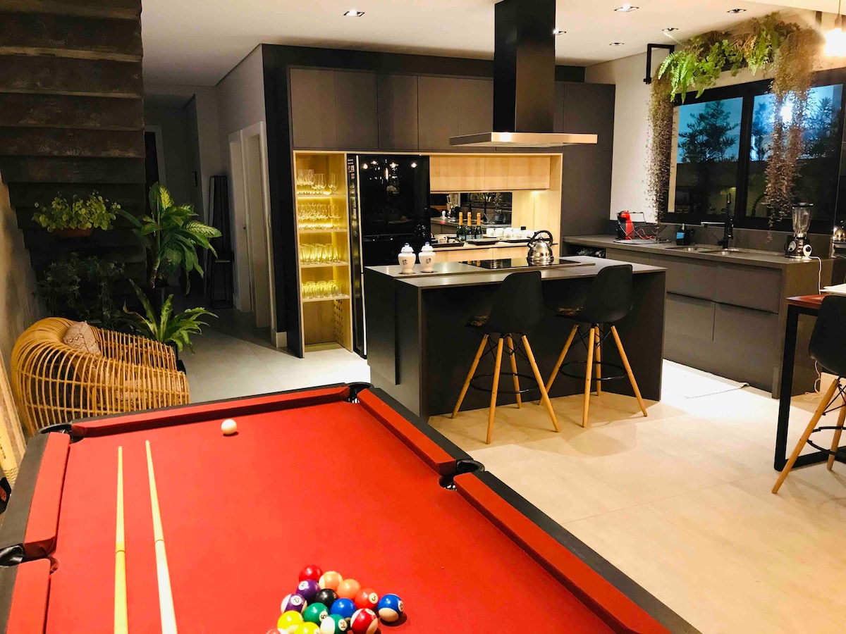 A modern living space is showcased, featuring a vibrant red pool table in the foreground. In the background, a sleek kitchen island is visible, accompanied by stylish stools. Natural light filters through a large window, illuminating the open layout with greenery adding a touch of nature.