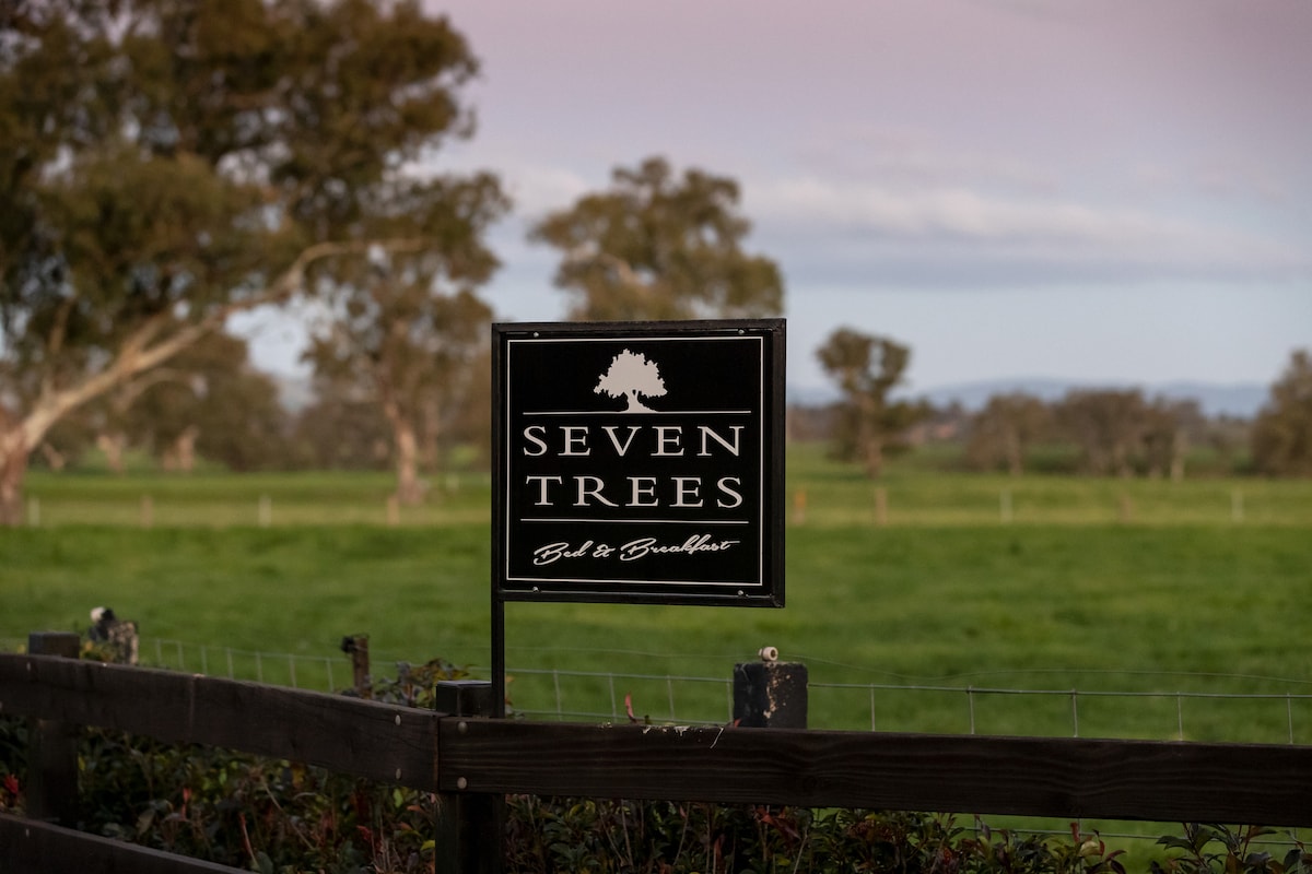 A sign displaying the name 'Seven Trees' is prominently positioned at the entrance. The background features a lush green pasture, bordered by trees and open fields, indicating a serene rural environment.