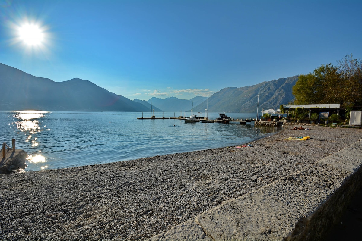 A calm waterfront scene features a pebbled beach extending towards a serene bay. Sunlight glimmers on the water, with mountains rising in the background. Several boats are moored at a dock, while a few sun loungers are positioned along the shore, providing spots for relaxation.