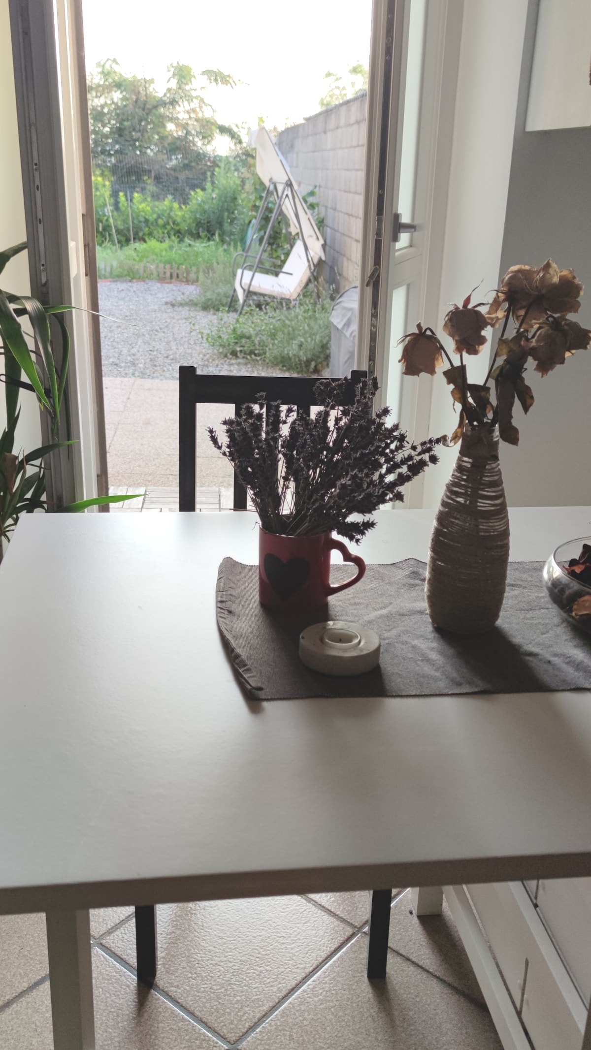 A dining table is set against a backdrop of an outdoor garden view, featuring a vase of dried flowers and a small cup. A candle rests on a dark table runner, while a chair enhances the dining area. Sunlight filters softly through the open door.