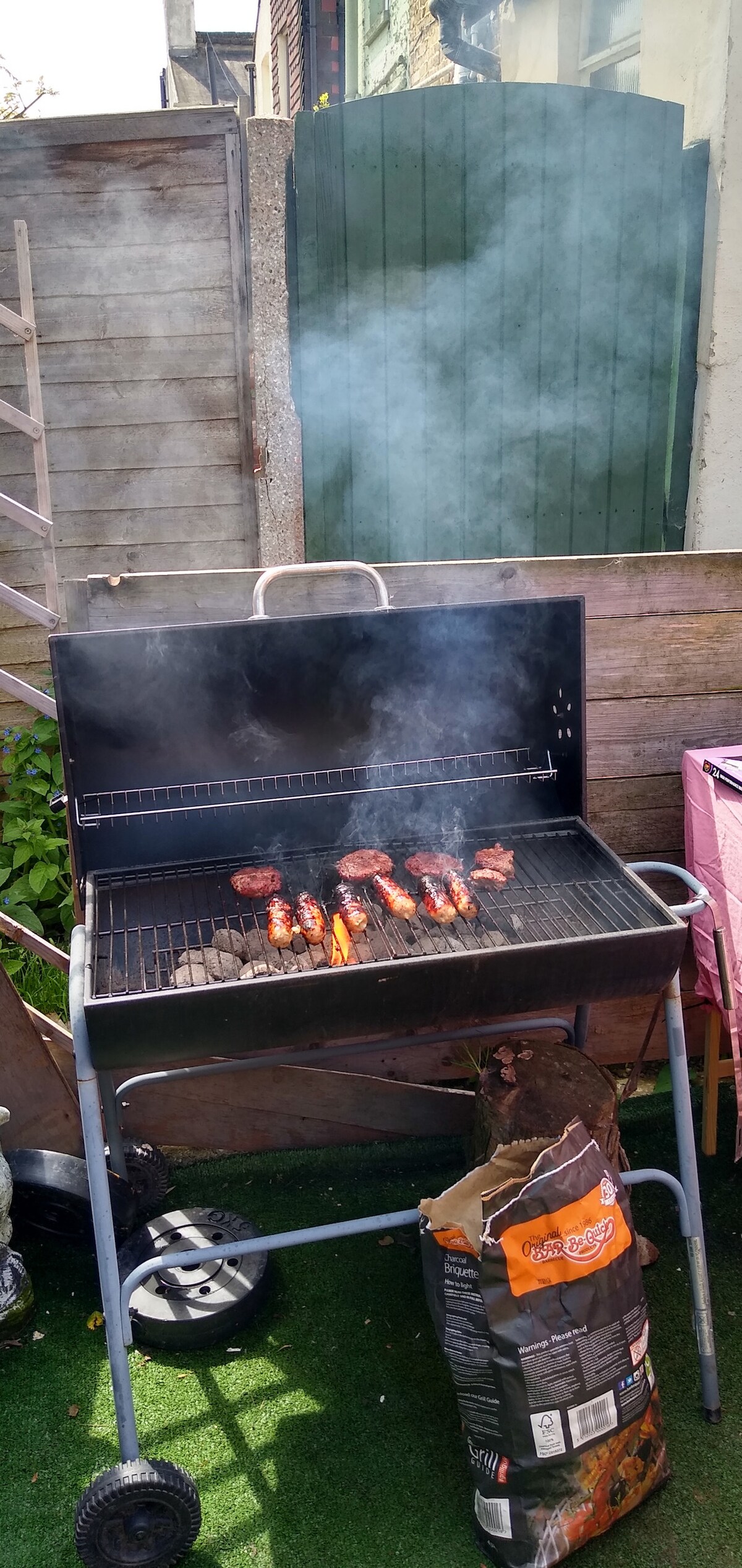A charcoal grill is seen in use, with several sausages and burgers cooking on the grates. Smoke rises gently from the grill, indicating active grilling. The green garden background includes a wooden fence and outdoor seating area, enhancing the outdoor ambiance.