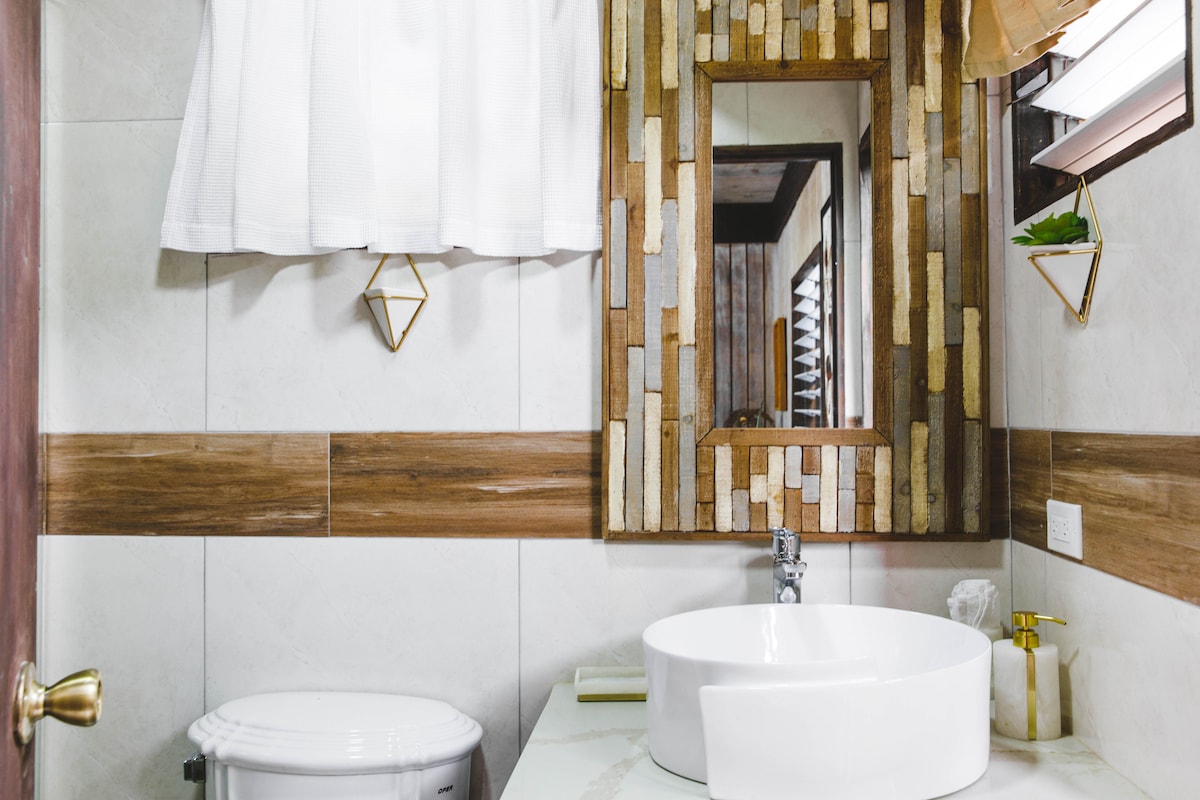 The bathroom features a round sink atop a marble countertop, accompanied by modern fixtures. A decorative mirror made of natural materials is mounted on the wall, enhancing the rustic style. Natural light filters through a window adorned with light fabric curtains.