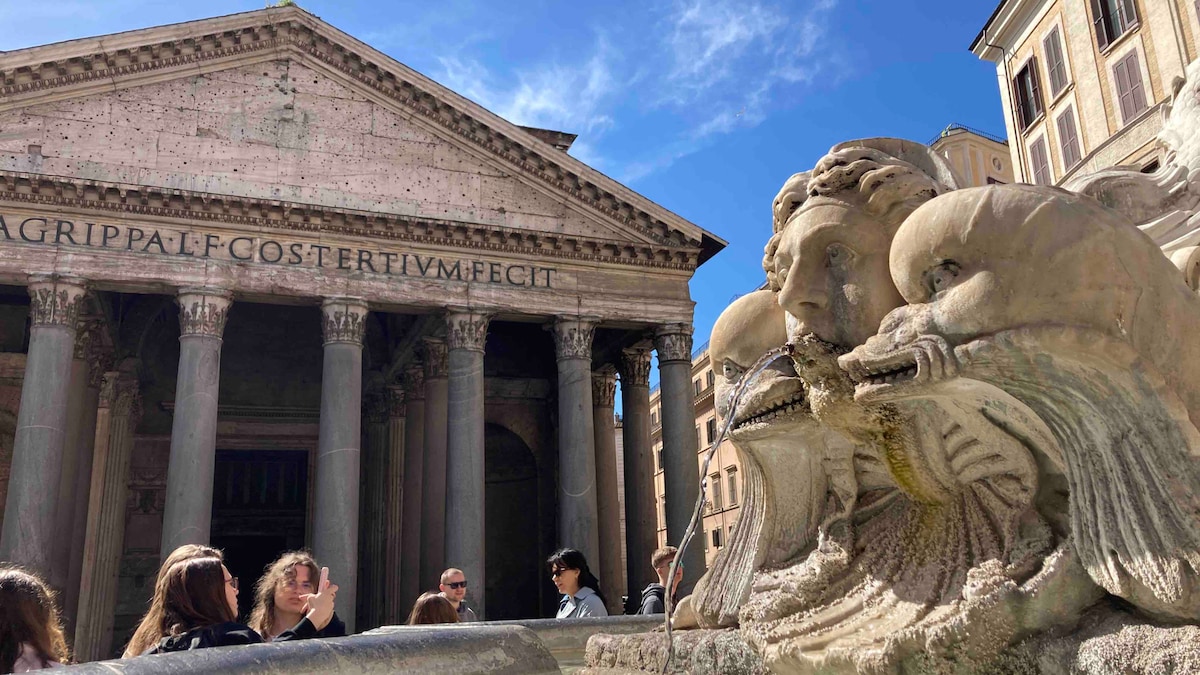 The iconic Pantheon is displayed in the background, showcasing its grand columns and classical architecture. A fountain with a sculptural feature occupies the foreground, creating a historical ambiance. Visitors can be seen enjoying the site under a clear blue sky.