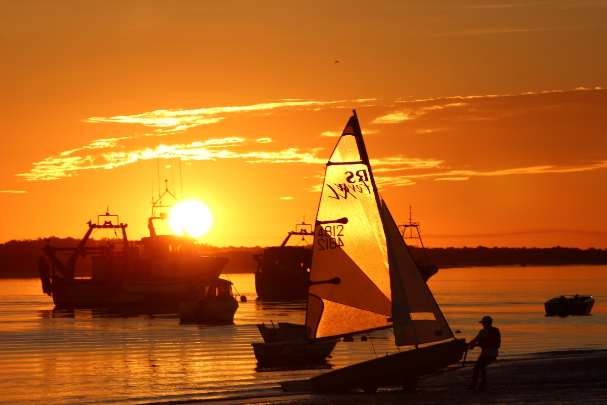 A vibrant sunset casts a golden hue over the water, with silhouettes of fishing boats in the background. A small sailboat is seen in the foreground, gliding across the smooth surface, creating a serene atmosphere that highlights the natural beauty of the river.
