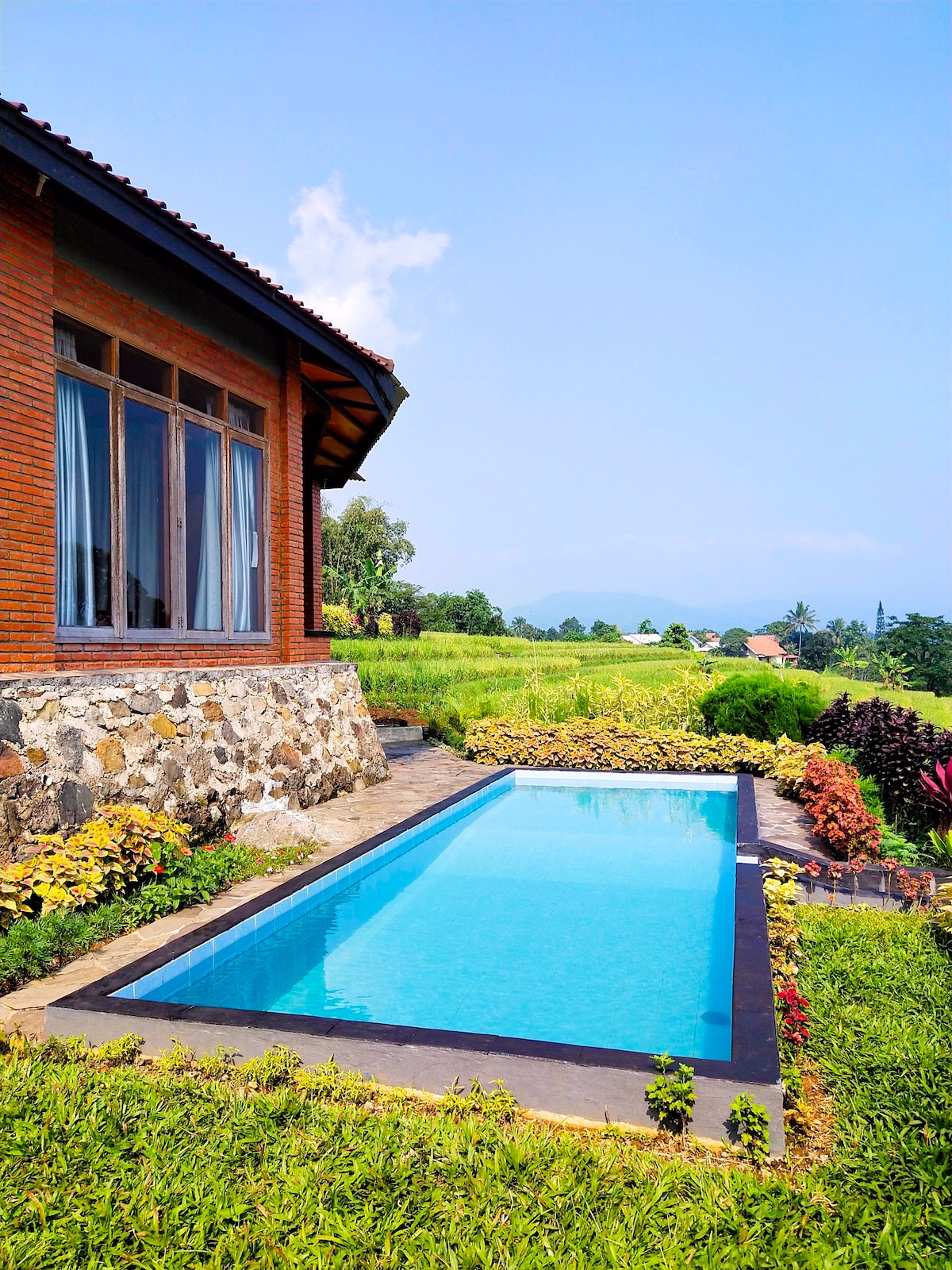 An inviting swimming pool is set beside a brick villa, surrounded by vibrant flower beds and lush grass. The clear blue water reflects the sky, while majestic mountains stand in the background, creating a serene outdoor space.