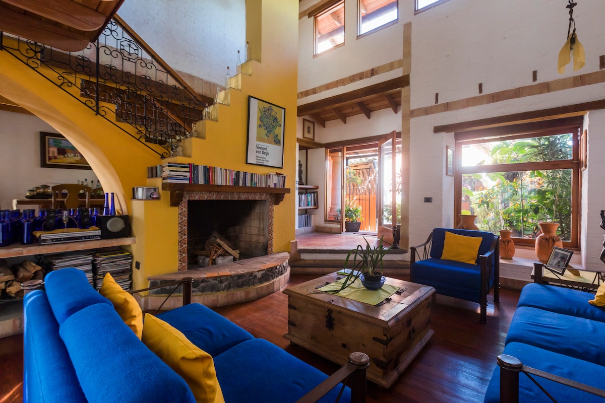 A bright living room features a double-height ceiling, with colorful blue sofas paired with yellow cushions. A rustic coffee table is centered atop wooden flooring. A stone fireplace adds warmth, while large windows invite natural light and offer views of the surrounding greenery.