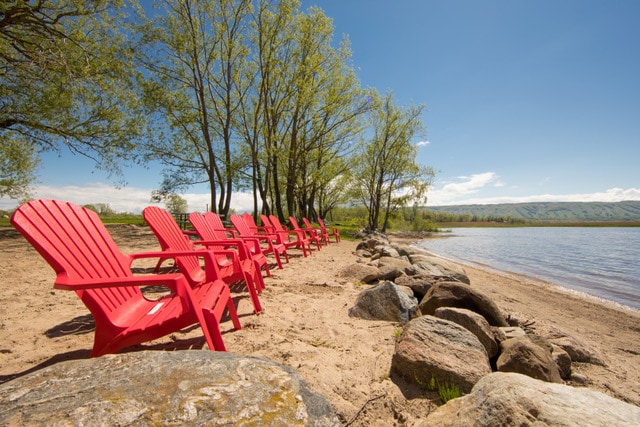 A row of vibrant red Adirondack chairs is positioned along a sandy beach, shaded by gentle trees. Smooth stones line the edge of the water, with a clear blue sky and distant hills visible in the background.