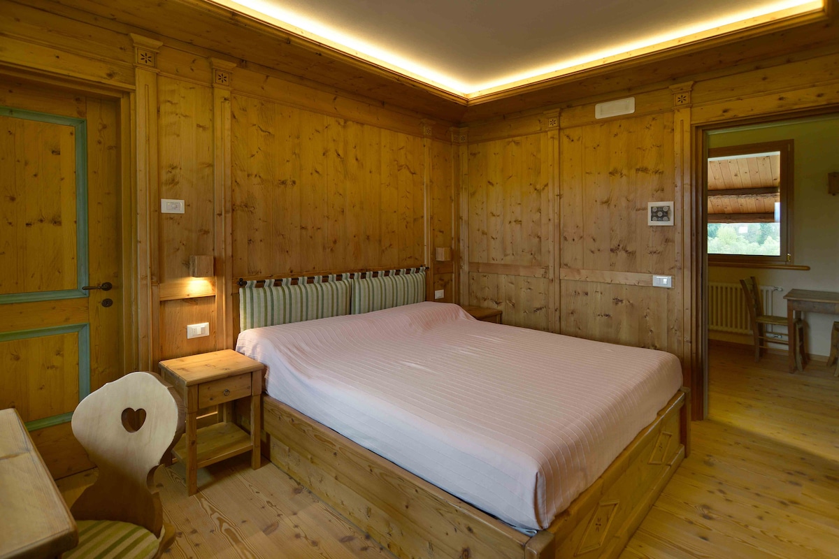 A spacious bedroom features warm wooden paneling, creating a cozy atmosphere. A large bed with a soft pink coverlet is positioned centrally, accompanied by a bedside table. A charming chair with a heart-shaped cutout adds character, while natural light filters through a window, enhancing the inviting environment.
