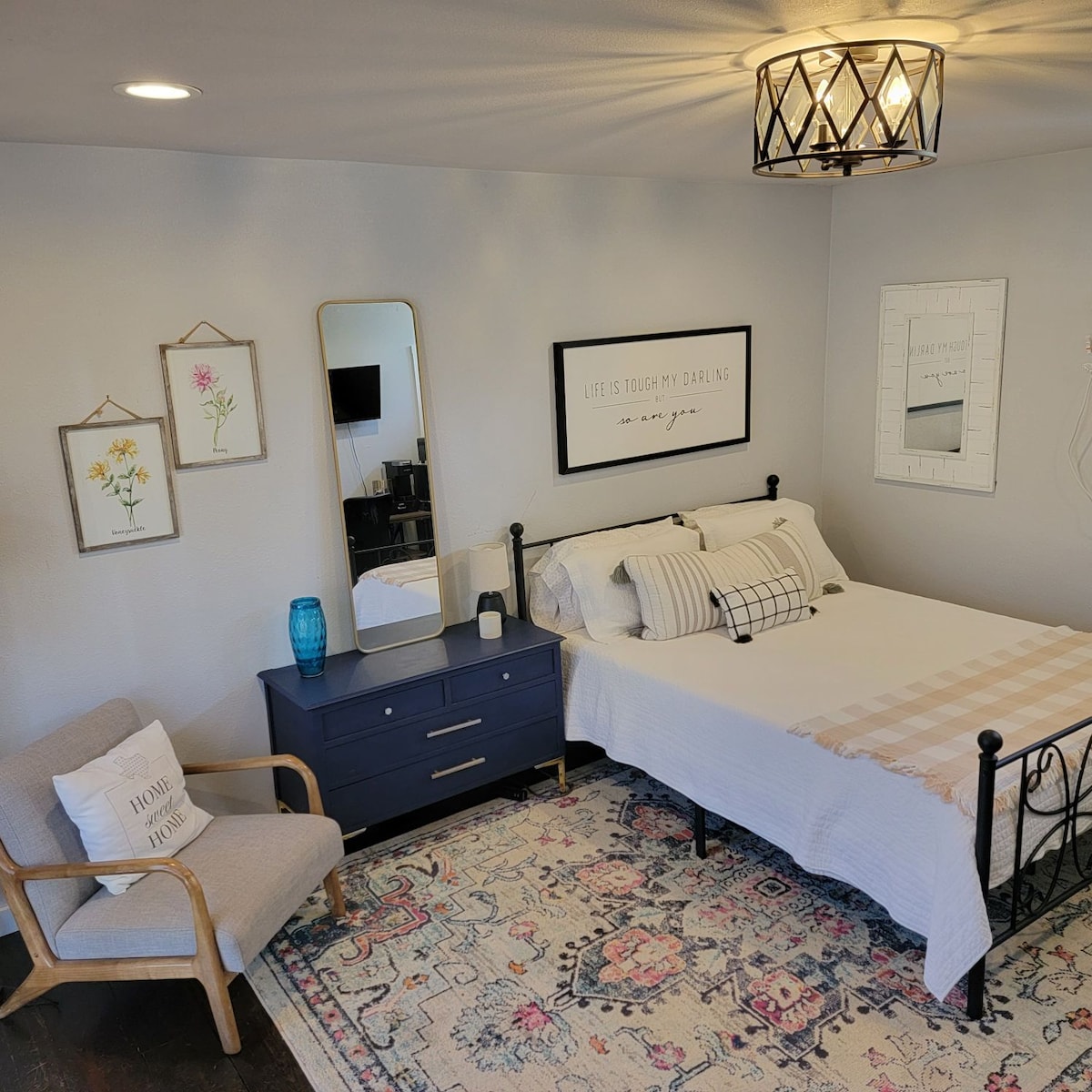 A well-organized bedroom features a spacious bed adorned with patterned pillows. A dresser in dark wood provides storage, while a mirror leans against the wall. Decorative wall art and a soft area rug add personal touches, and a comfortable chair is placed in a corner.