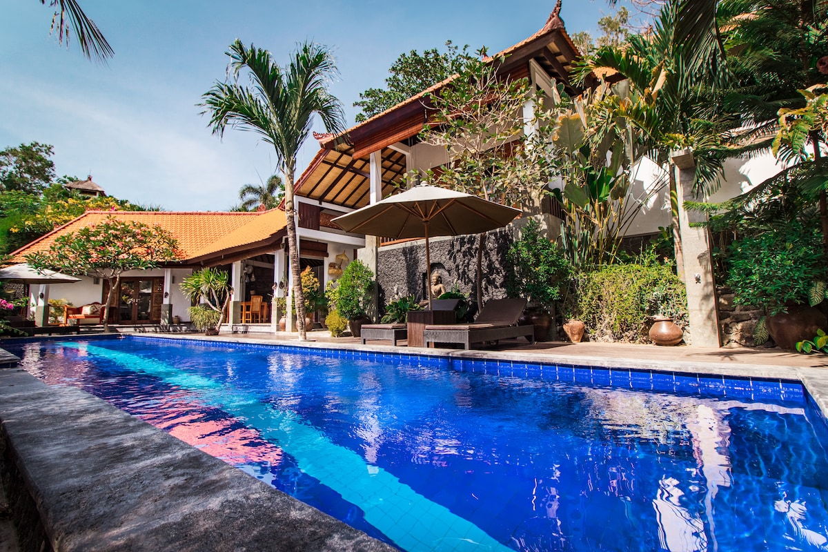 A serene outdoor area displays a vibrant blue swimming pool surrounded by tropical plants. Lounge chairs are positioned under a large umbrella, with a traditional villa and a covered seating area seen in the background beneath a tiled roof.