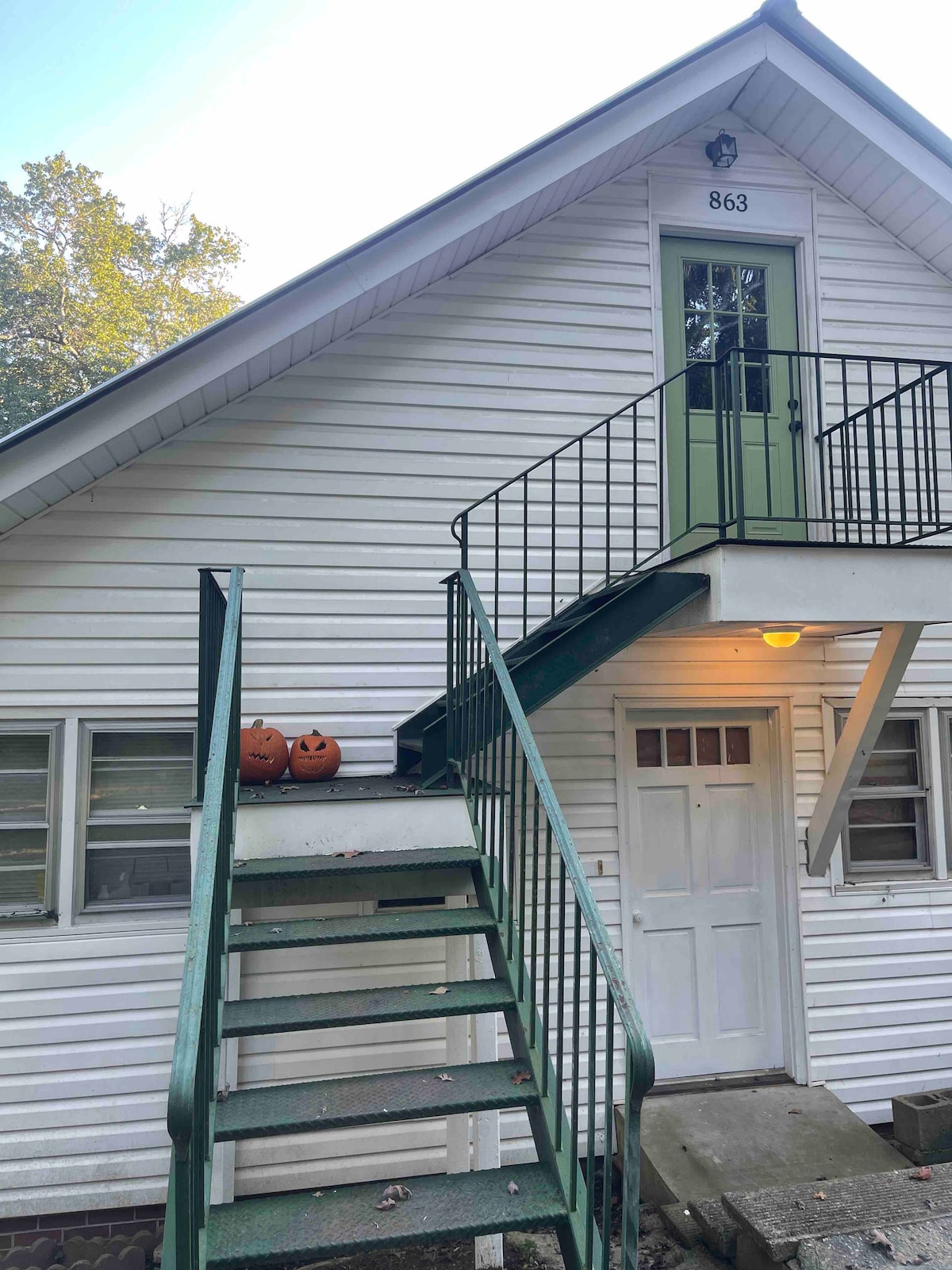 The exterior of a two-story structure is shown, featuring a white staircase with a green railing leading to the upper level. Two carved pumpkins rest at the base of the stairs, and light filters through the windows of the building.