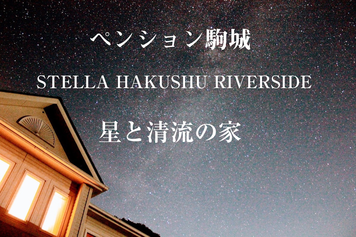 A night sky filled with stars provides a serene backdrop to the building, which features prominent architectural lines and warm lighting from the windows. The name 'Pension Komaki' is elegantly displayed, along with 'Stella Hakushu Riverside' in both Japanese and English.