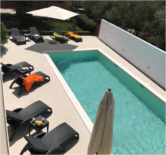 A clear swimming pool is bordered by a spacious deck that includes several lounge chairs. An umbrella provides shade for the seating area. Brightly colored towels and fruit on a table enhance the relaxation space, surrounded by greenery for a serene setting.