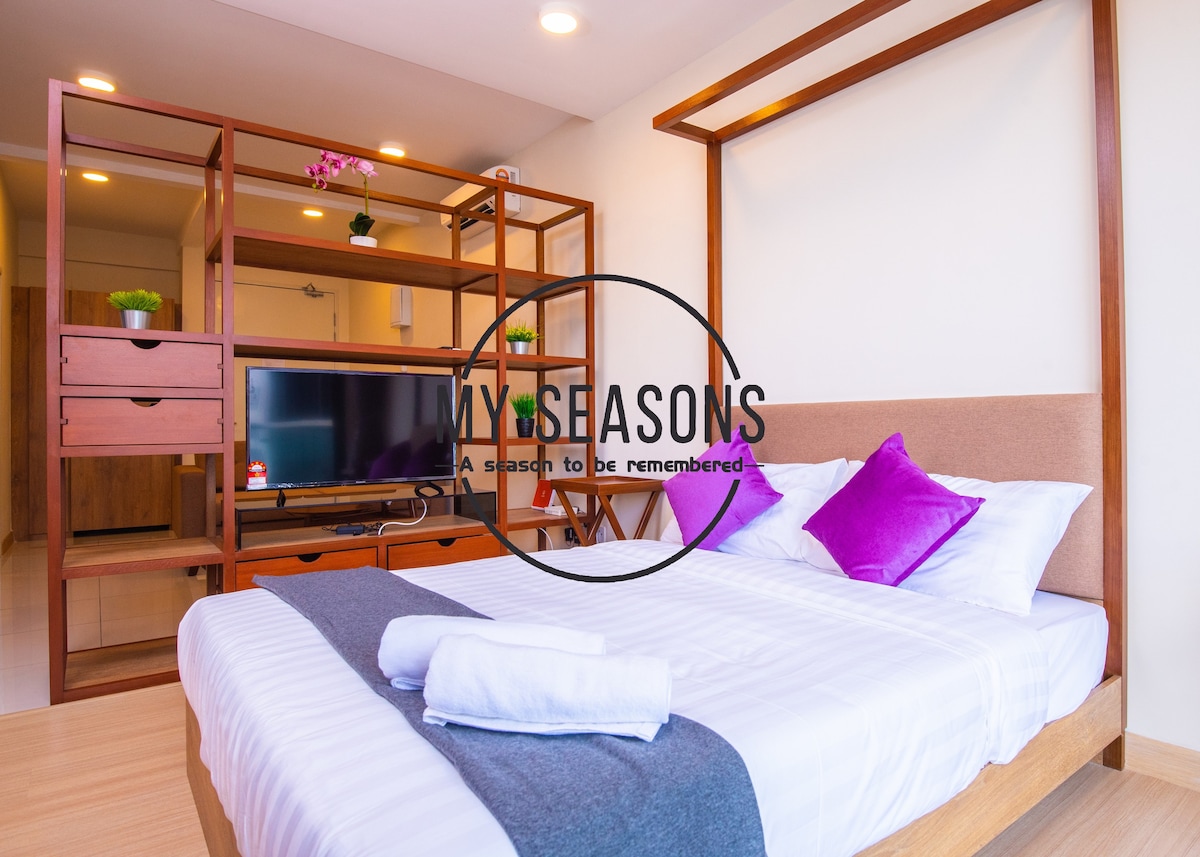 A spacious sleeping area features a queen-sized bed dressed in fresh white linens and accented by purple pillows. A wooden shelving unit with decorative plants and a television adds functionality. Soft lighting enhances the inviting ambiance for relaxation.