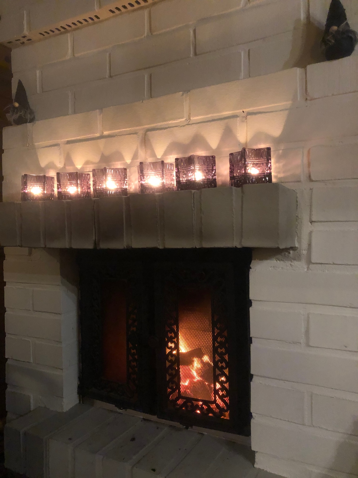 A brick fireplace is visible, featuring a metallic grill front that encloses a warm glow from the fire inside. Six candle holders with soft, flickering lights are arranged along the mantel above, creating a cozy atmosphere.