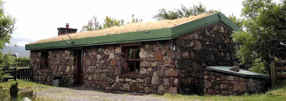 A stone cottage is featured, showcasing natural materials and a green grass-covered roof. The structure stands amidst greenery, with a single window offering a view of the surroundings. The rustic appearance is complemented by simple landscaping, adding charm to the remote setting.