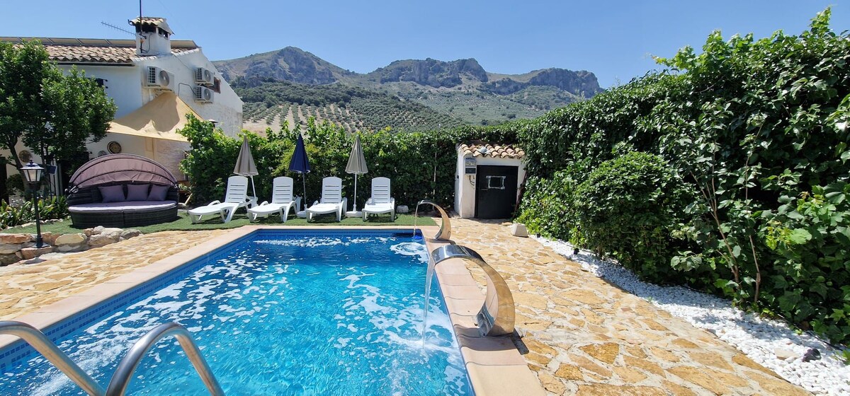 A refreshing swimming pool is featured, surrounded by lounge chairs under the sun. Lush greenery lines the area, and scenic mountains are visible in the background. A stone pathway leads to the pool, enhancing the outdoor space.
