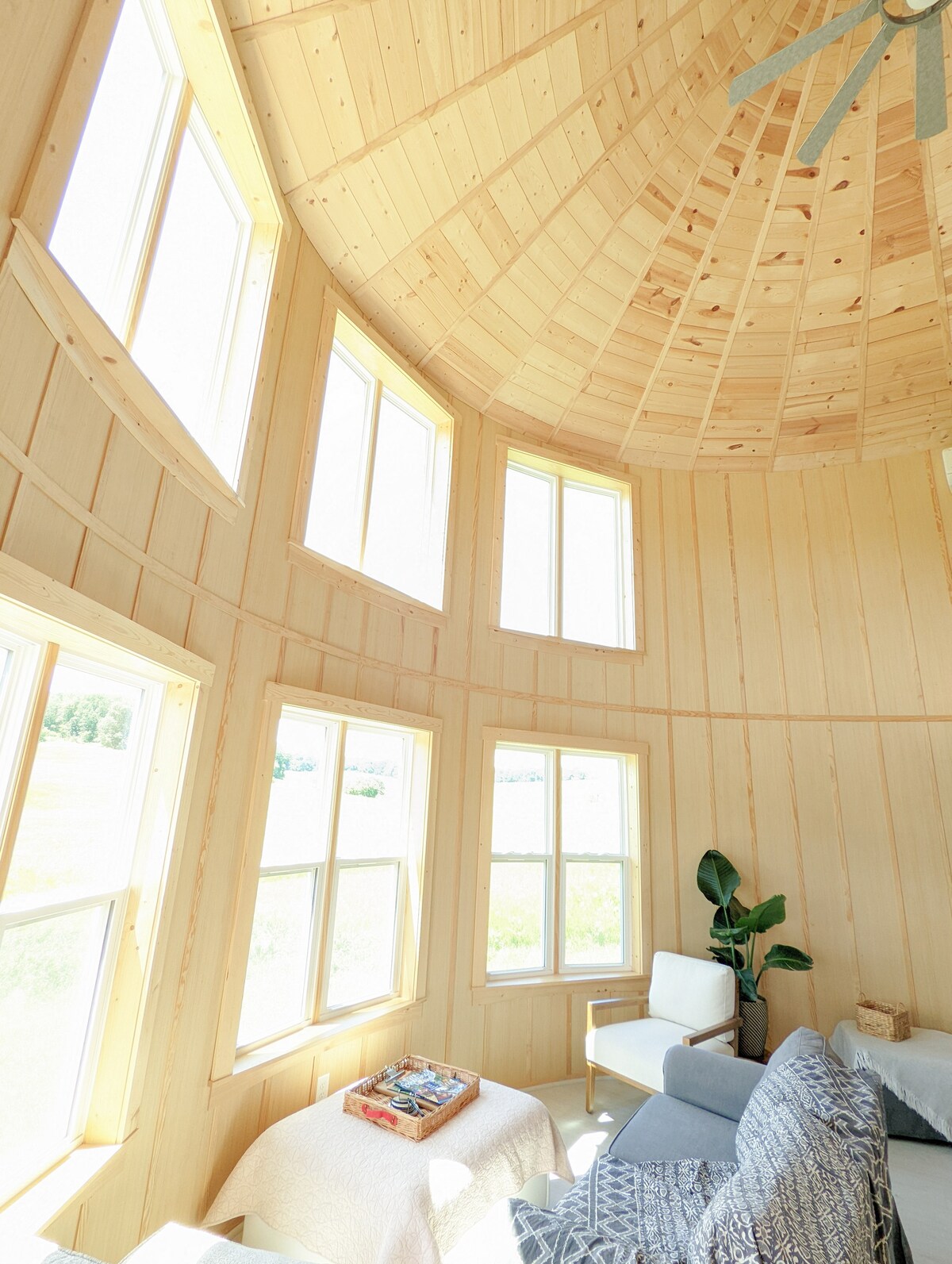 The interior of the cabin features a circular design with multiple floor-to-ceiling windows, allowing abundant natural light. Light wood paneling creates a warm atmosphere, while a cozy seating area includes a sofa, patterned throw, and a small coffee table displaying reading materials.
