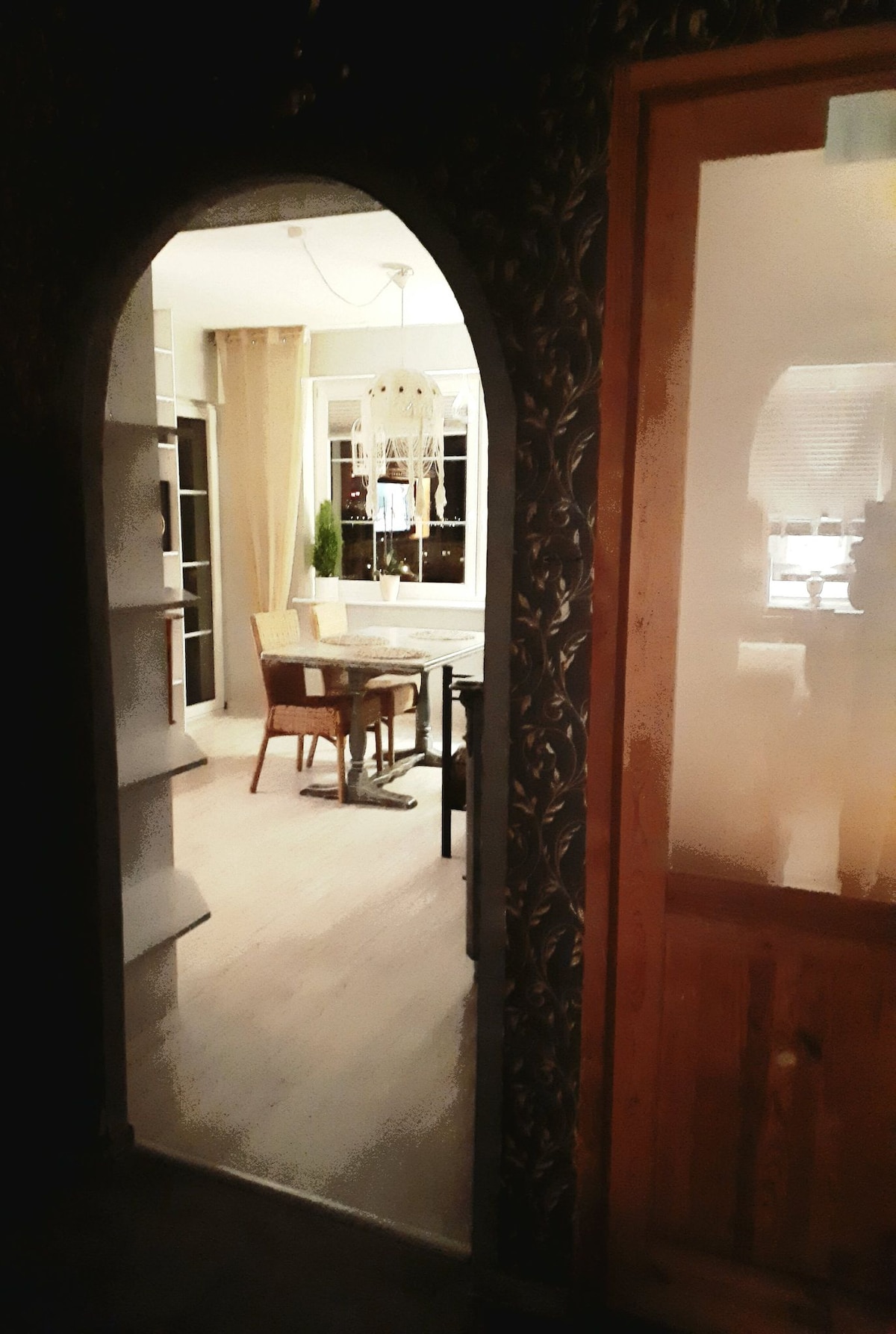 A view through a curved archway reveals a dining area with a small table and chairs under a pendant light. Light-colored curtains frame the windows, contributing to a bright atmosphere. Shelving is visible along the wall, enhancing the space's charm.