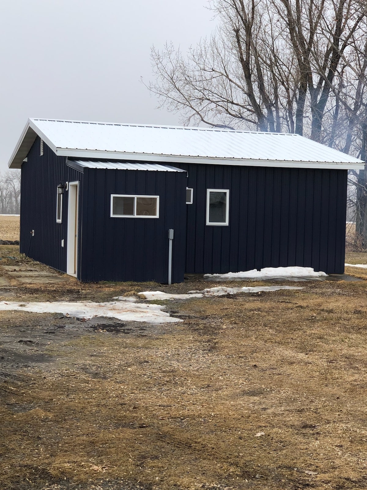 A small, modern cabin is seen with dark exterior siding and a sloped roof. The building features a window design with two small windows on one side and a door entrance. Surrounding the cabin, patches of snow and bare ground are visible, indicating early spring.
