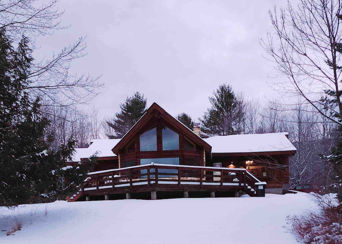 A spacious log home is set against a snowy landscape, featuring a prominent front deck with railings, inviting entry. Large windows provide an unobstructed view of the surrounding wooded area, while tall trees stand nearby, enhancing the natural surroundings.