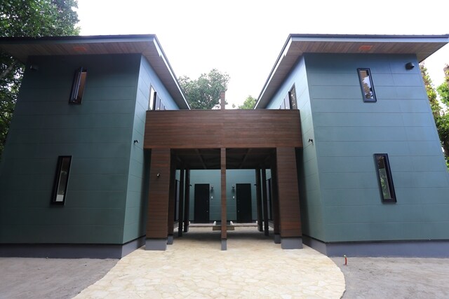 The exterior of a two-story vacation rental is shown, featuring a modern design with teal walls and a wooden overhang. Symmetrical structures are framed by stone pathways and greenery, with tall windows allowing natural light inside.