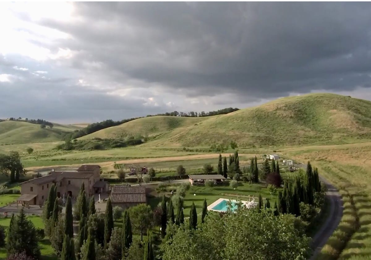 An aerial view captures the expansive Tuscan landscape, featuring gently rolling hills and a charming countryside estate. The property includes lush green gardens, a swimming pool, and neatly arranged trees, all set against a moody sky, enhancing the serene atmosphere of the surroundings.