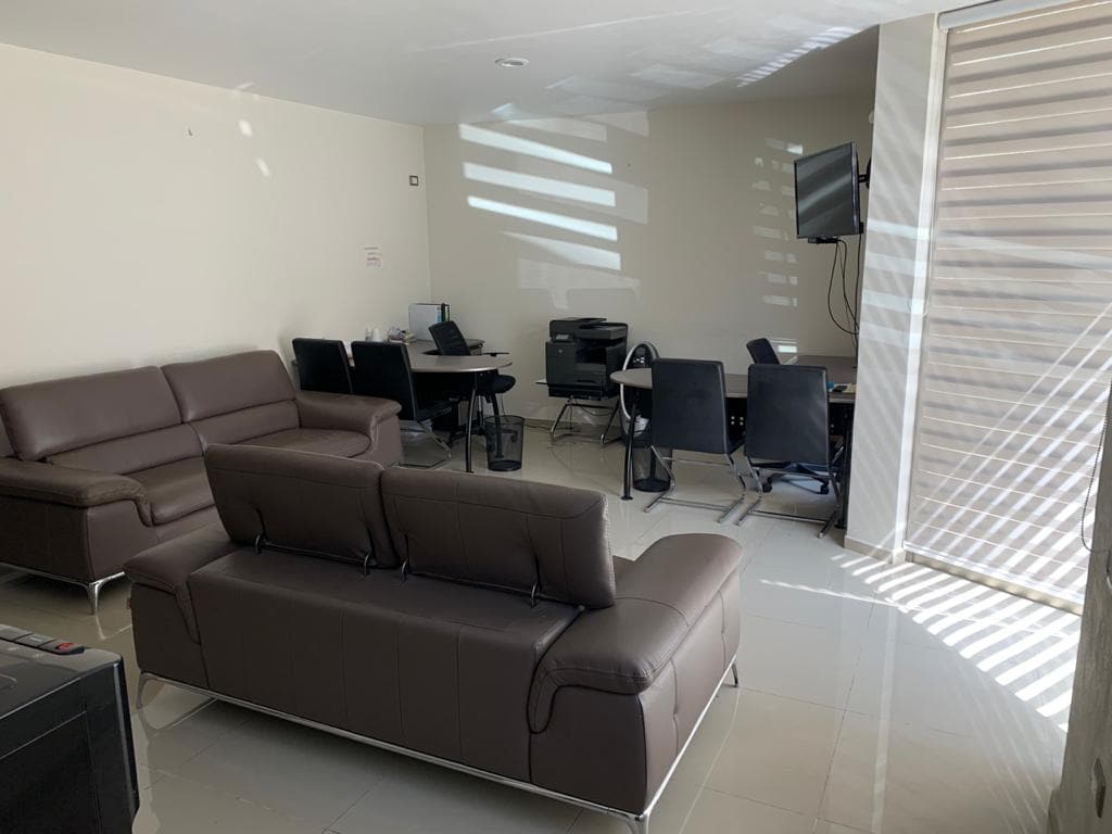 A modern living area is arranged with a brown leather sofa and several black chairs around a desk. Natural light filters through horizontal blinds, casting soft shadows on the walls. A wall-mounted television is visible, enhancing the multifunctional space.