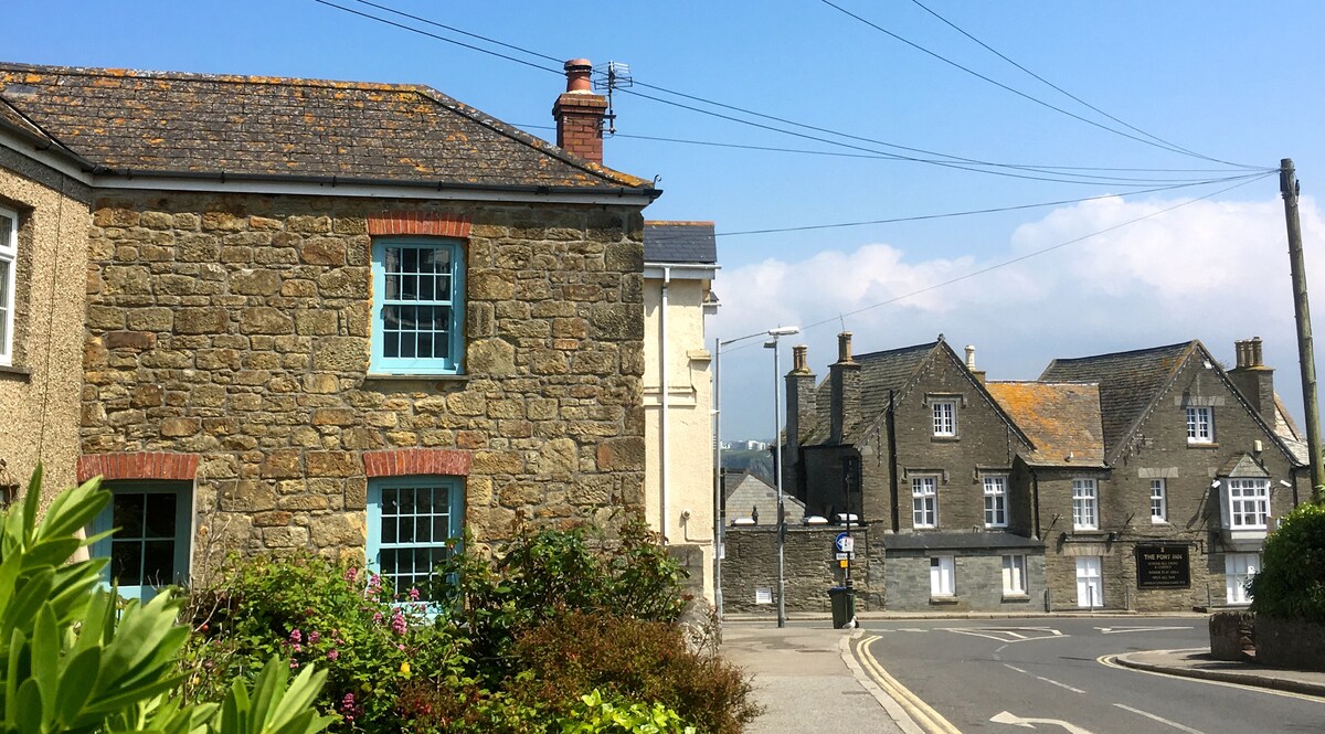 The exterior of a charming stone cottage is visible, featuring quaint blue windows that complement the rustic appeal. Nearby buildings, with varied architecture, frame the street, while greenery adds a touch of natural beauty to the scene.
