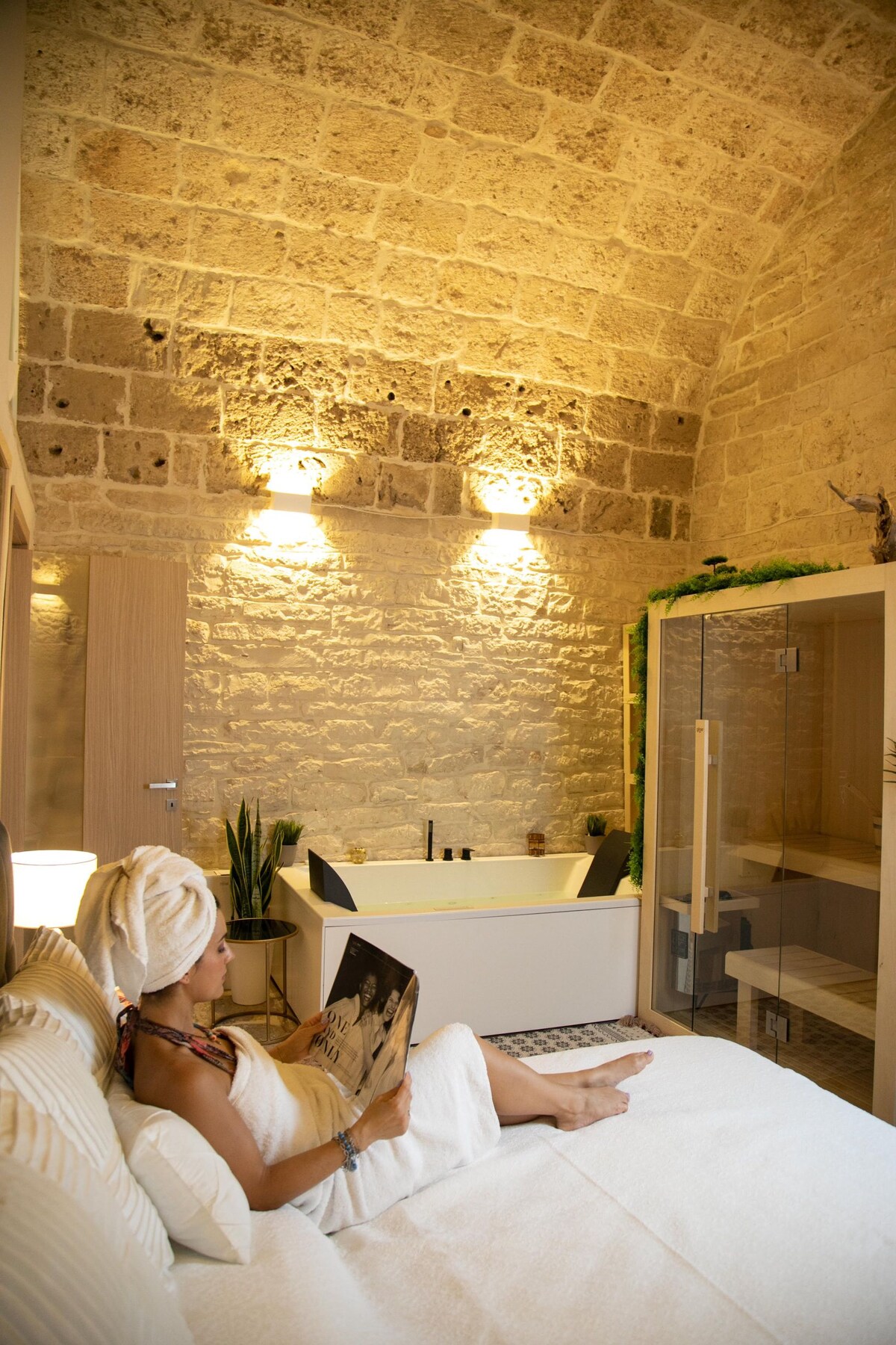 A serene bathroom setting is captured, featuring a spacious jacuzzi tub next to a glass shower enclosure. Natural stone walls are highlighted by soft lighting. A plush bed is visible, accompanied by a person relaxing and reading, adding to the tranquility of the space.
