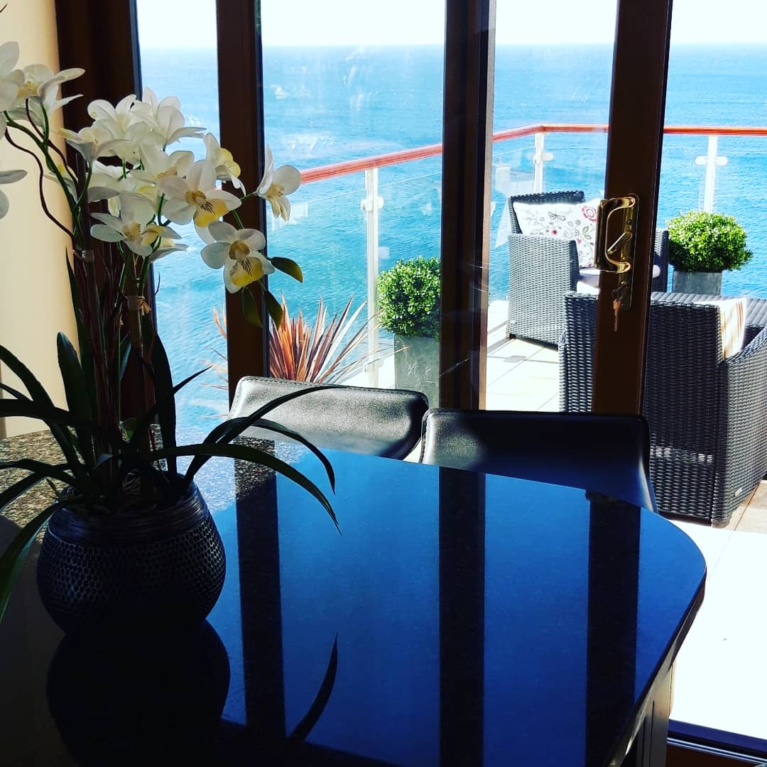 A well-furnished seating area is visible through large glass doors, offering views of the sea. A dark table reflects sunlight, while an elegant vase with white orchids adds a touch of greenery. On the balcony, outdoor seating arrangements can be seen against the backdrop of water.