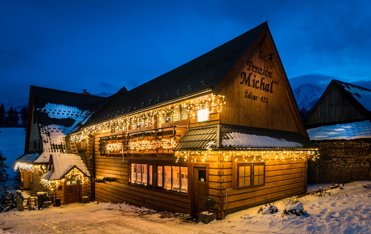The charming wooden exterior of the chalet is adorned with warm white string lights, creating a welcoming glow against the evening sky. Snow blankets the ground, while the roof features classic wooden accents. Nearby structures, partially obscured by snow, blend into the serene winter landscape.