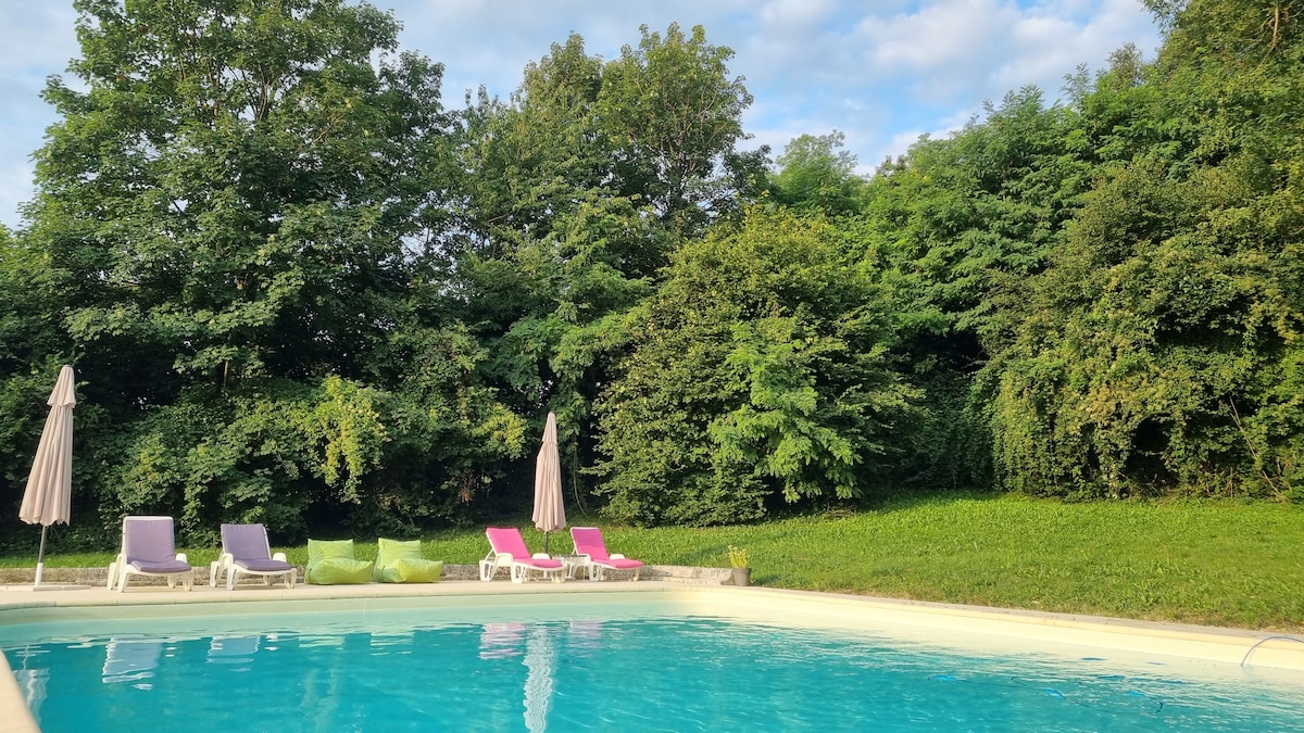 A clear swimming pool is surrounded by lush greenery, including tall trees and shrubs. Several pool loungers in soft pastels are positioned nearby, with two umbrellas providing shade. The inviting water reflects the blue sky, creating a peaceful outdoor space.