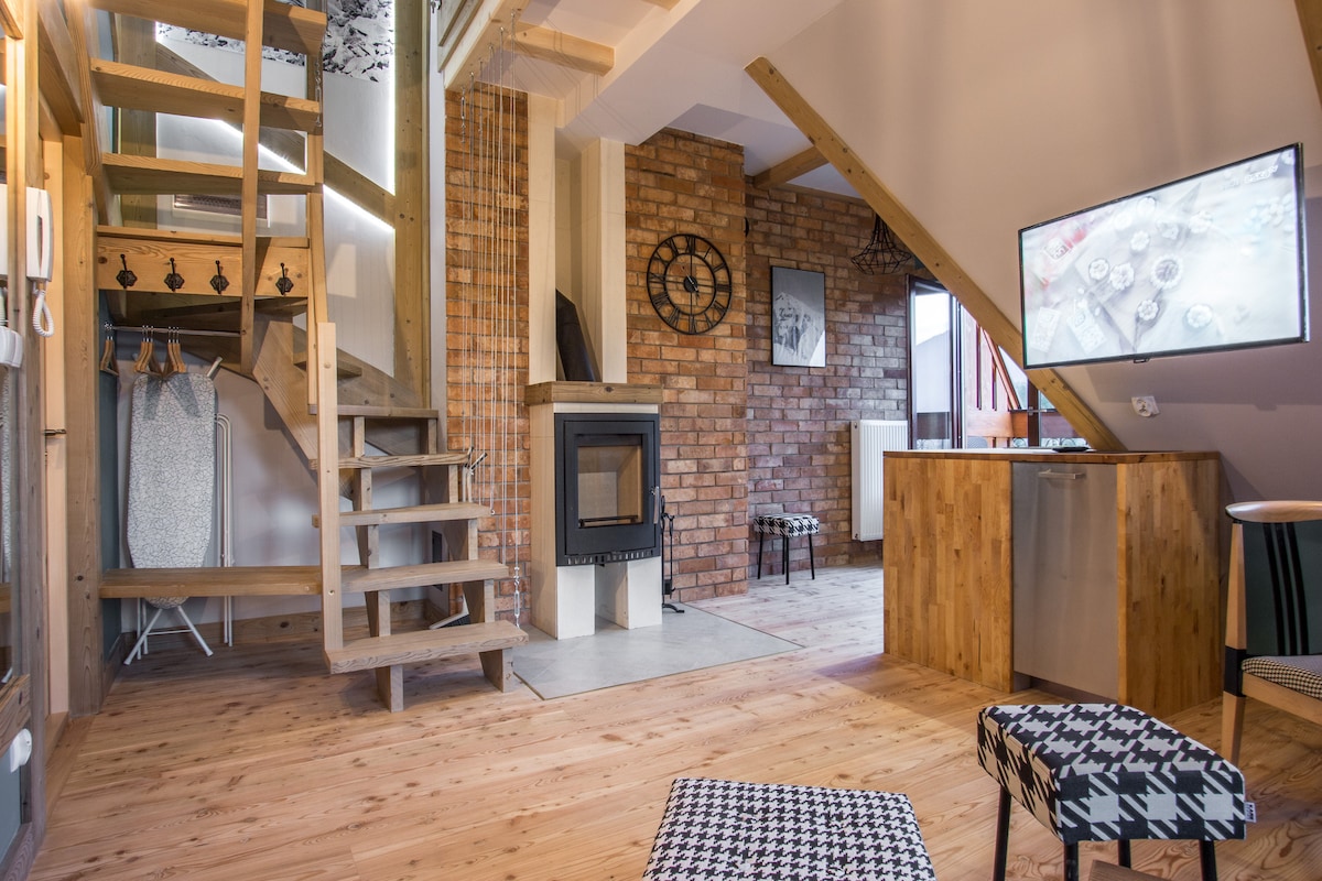 The interior showcases a blend of wood and brick elements, creating a warm atmosphere. A corner fireplace is visible, surrounded by sleek furniture. A staircase leads to the upper level, and a television is mounted on the wall, complementing the open space.